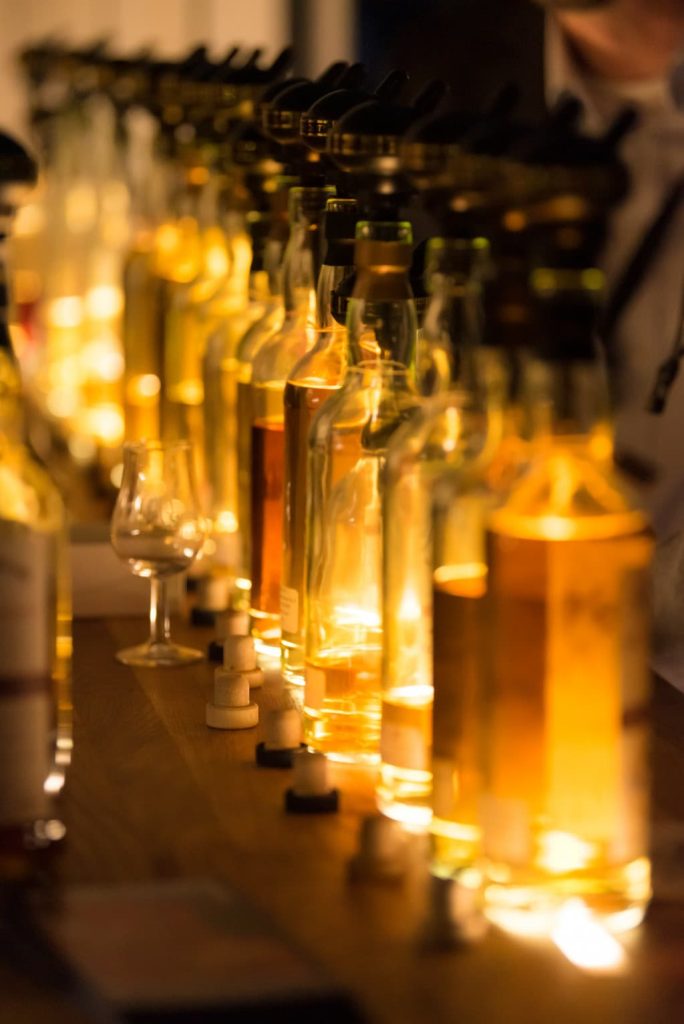 Bernsteinfarben-leuchtende Flaschen sind in einer Reiher auf einer einer Theke aufgereiht. Vor ihnen steht ein kleines Glas mit Stil.